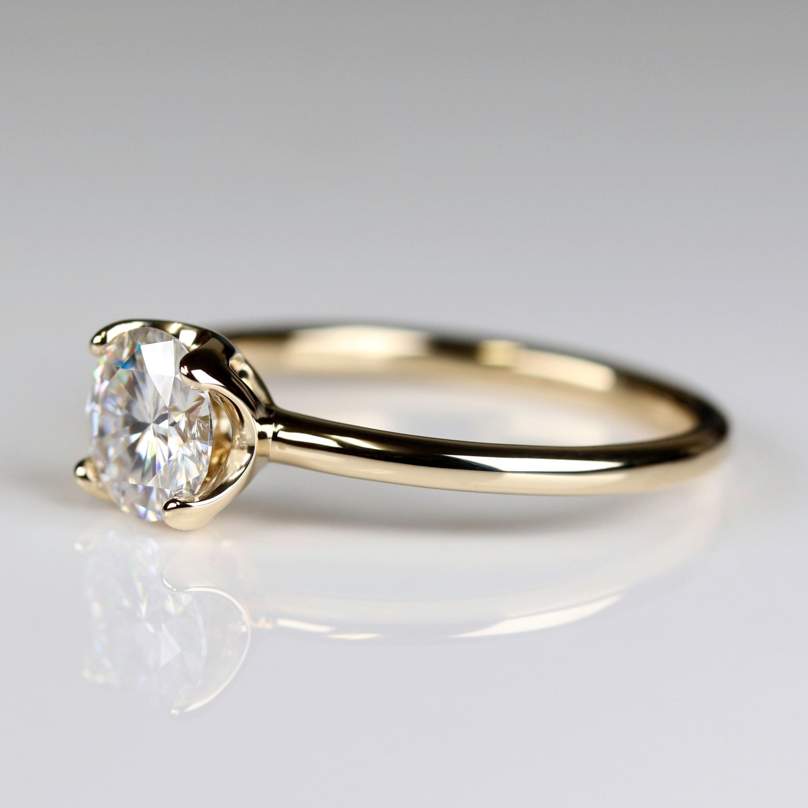 Side angle of a yellow gold solitaire engagement ring with a prong-set white moissanite. The center stone is around 6mm in diameter, and is set in a tulip setting.