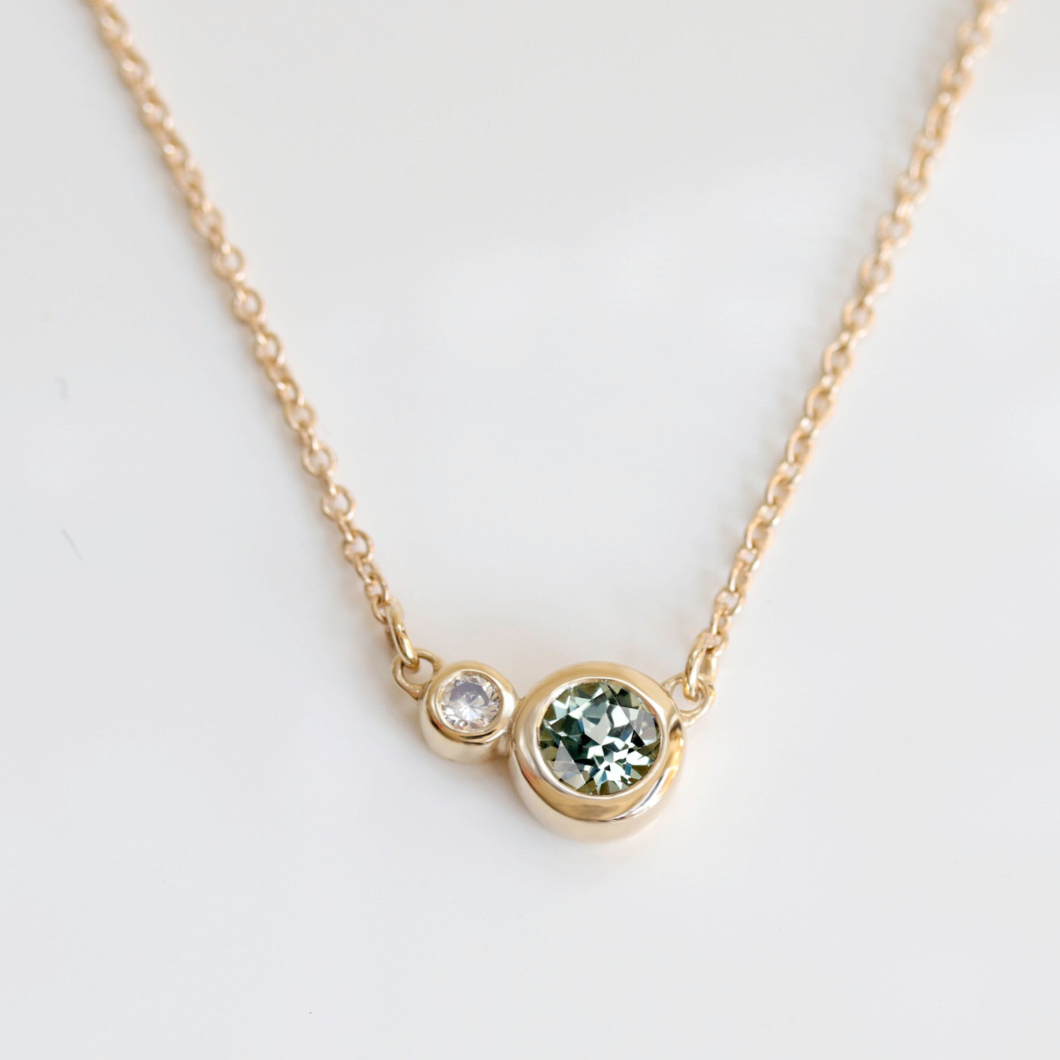 A fixed pendant necklace in 14k yellow gold. A light green lab-grown sapphire and white diamond are bezel set.
