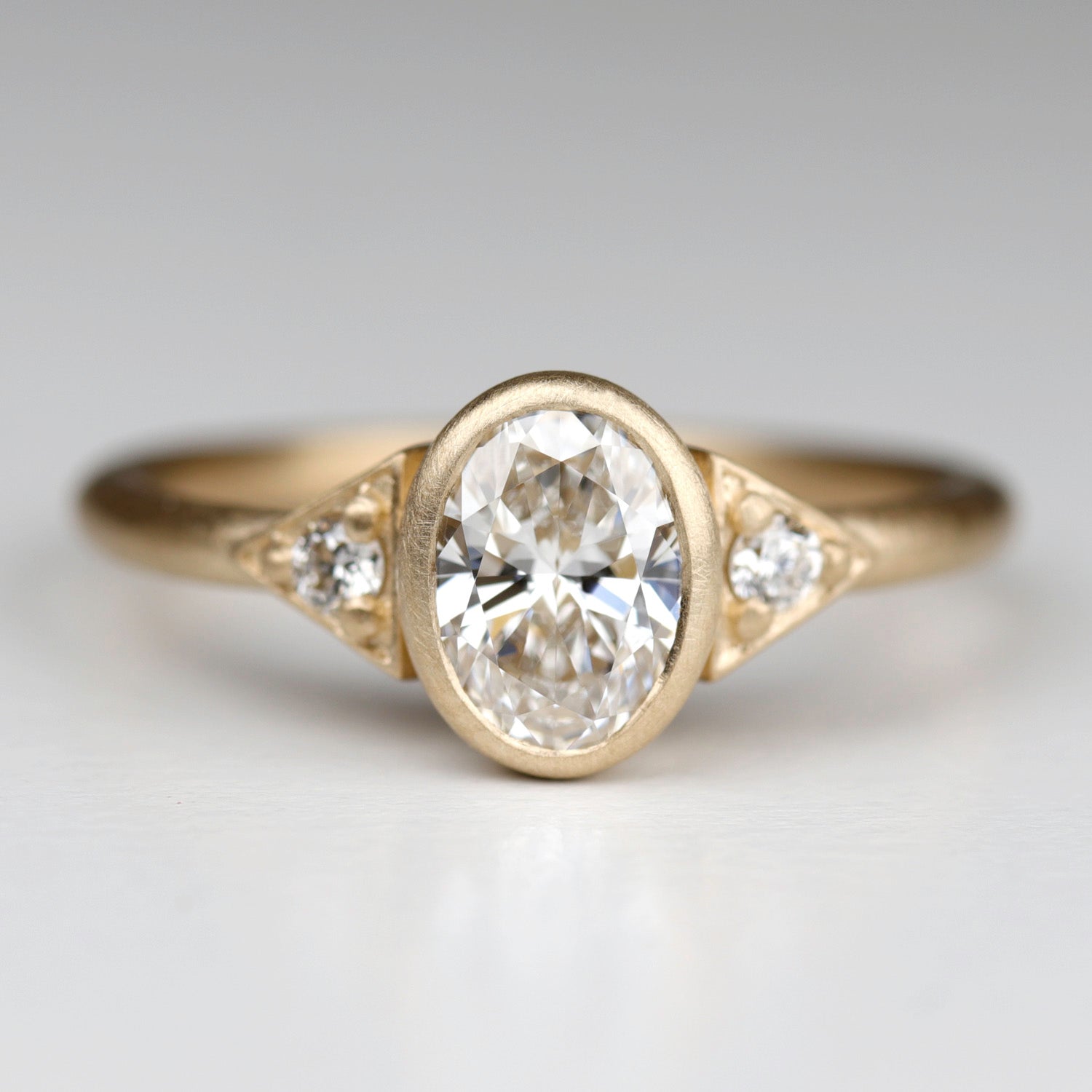 A three stone oval engagement ring in matte yellow gold with diamonds. The center stone is bezel-set and the round accent stones have prongs.