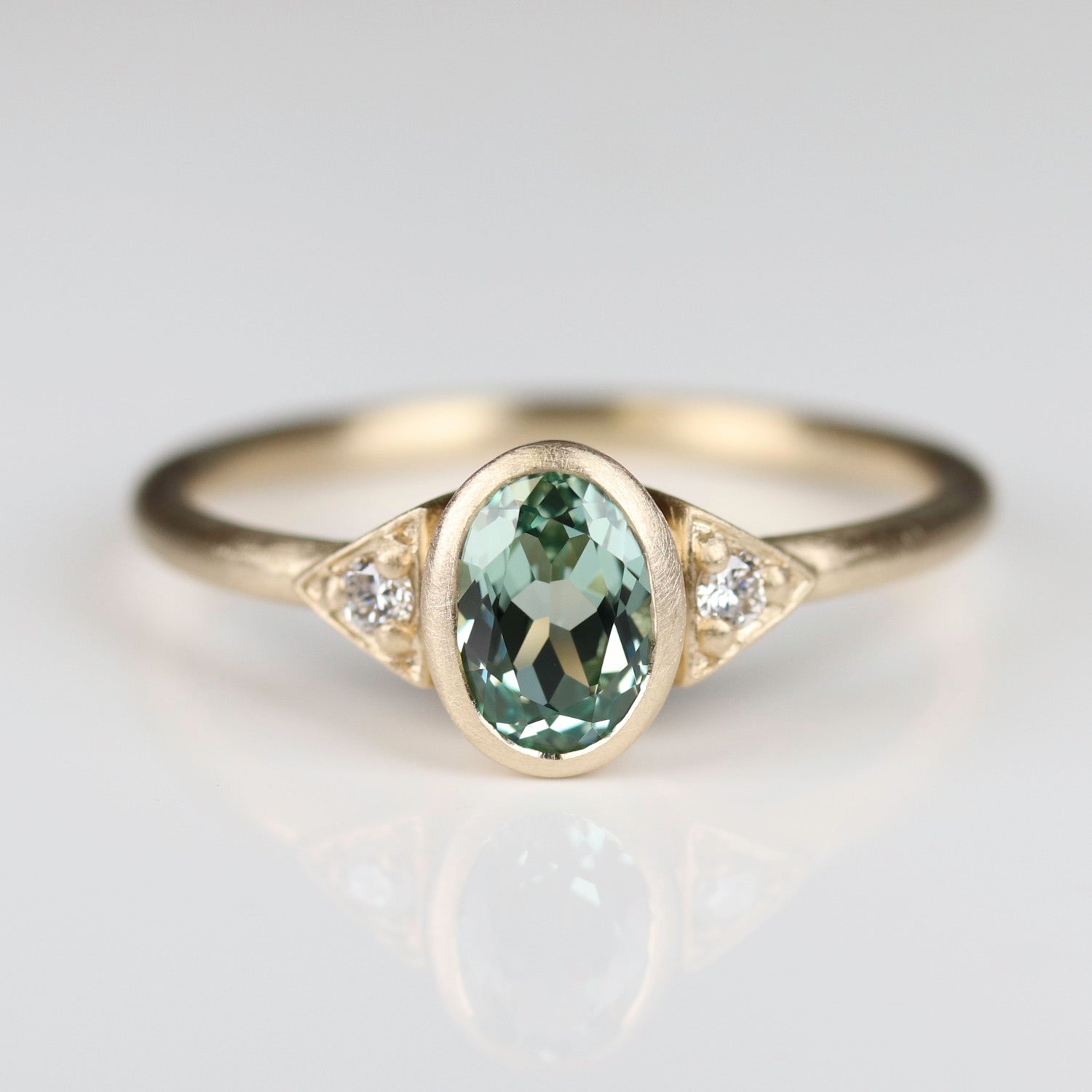 A three stone oval engagement ring in matte yellow gold with a lab-grown light green sapphire and accent diamonds. The center stone is bezel-set and the round accent stones have prongs.
