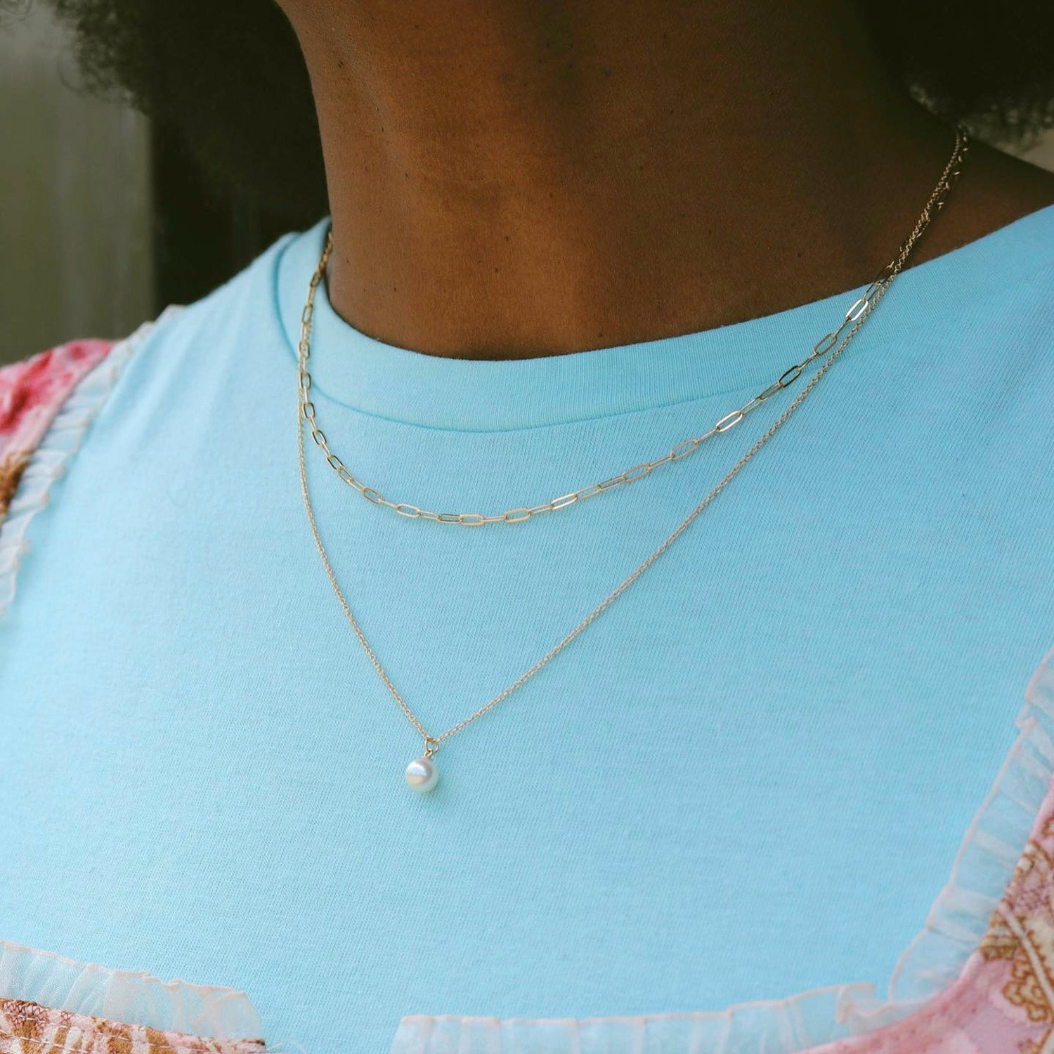 Model wearing a pearl drop pendant hanging from a simple chain. The necklace is layered with a paperclip chain.