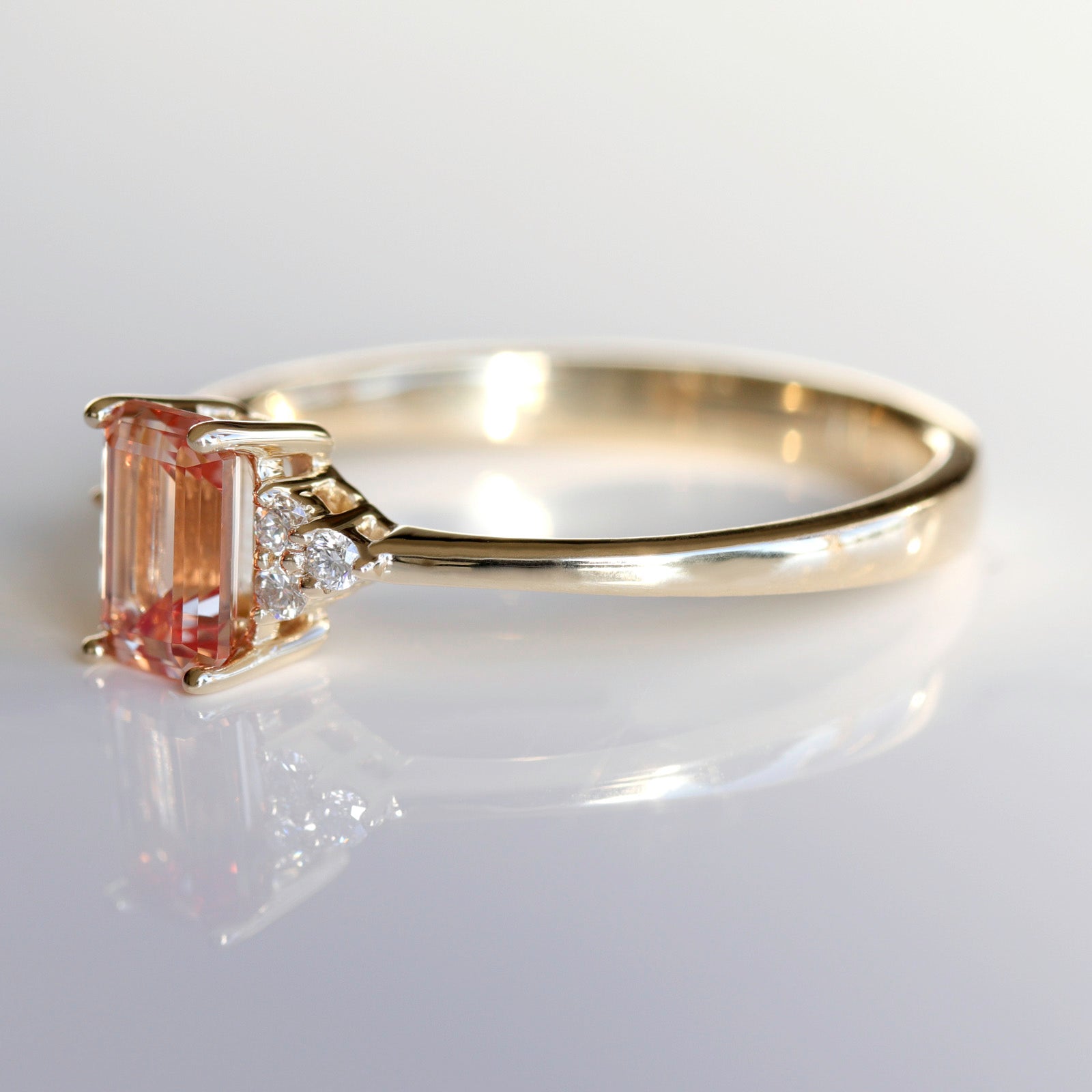Side angle of a seven stone cluster engagement ring style in shiny yellow gold. The ring has a peach champagne sapphire center stone in an emerald cut.