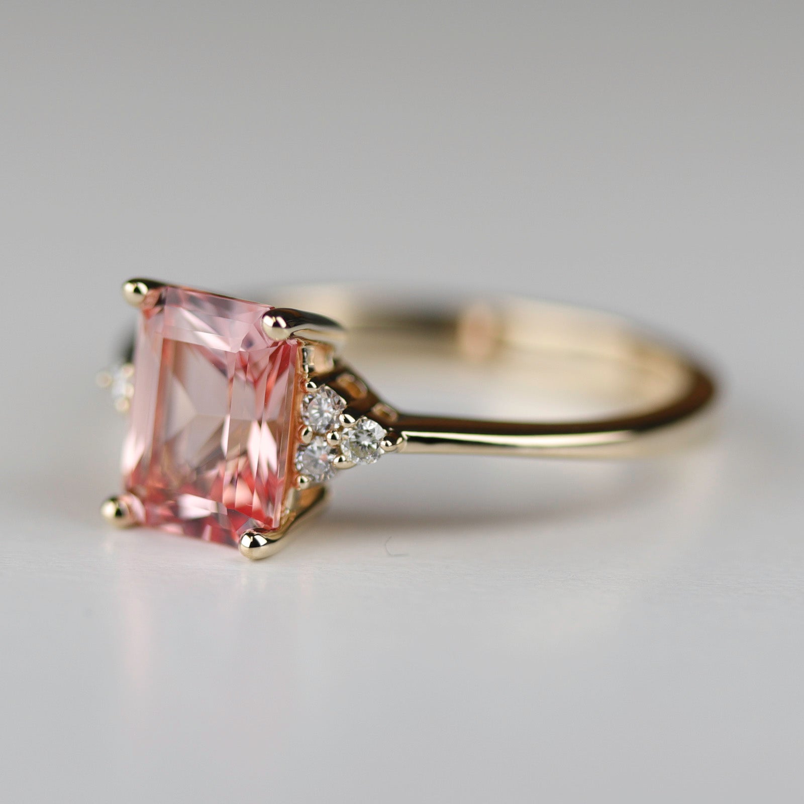 Side angle of a seven stone cluster engagement ring style in shiny yellow gold. The ring has a peach champagne sapphire center stone in an emerald cut.