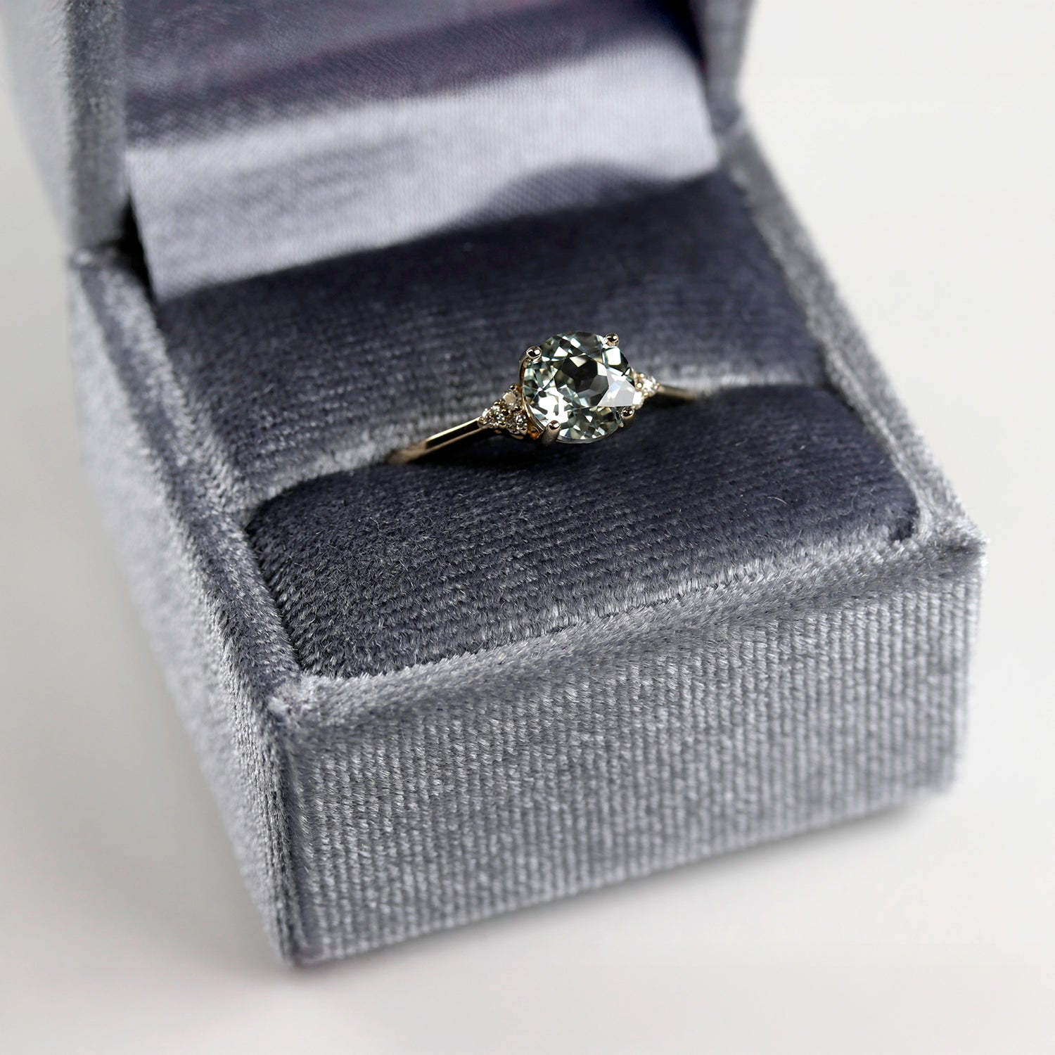 A sapphire engagement ring in a silver gray velvet clamshell ring box.