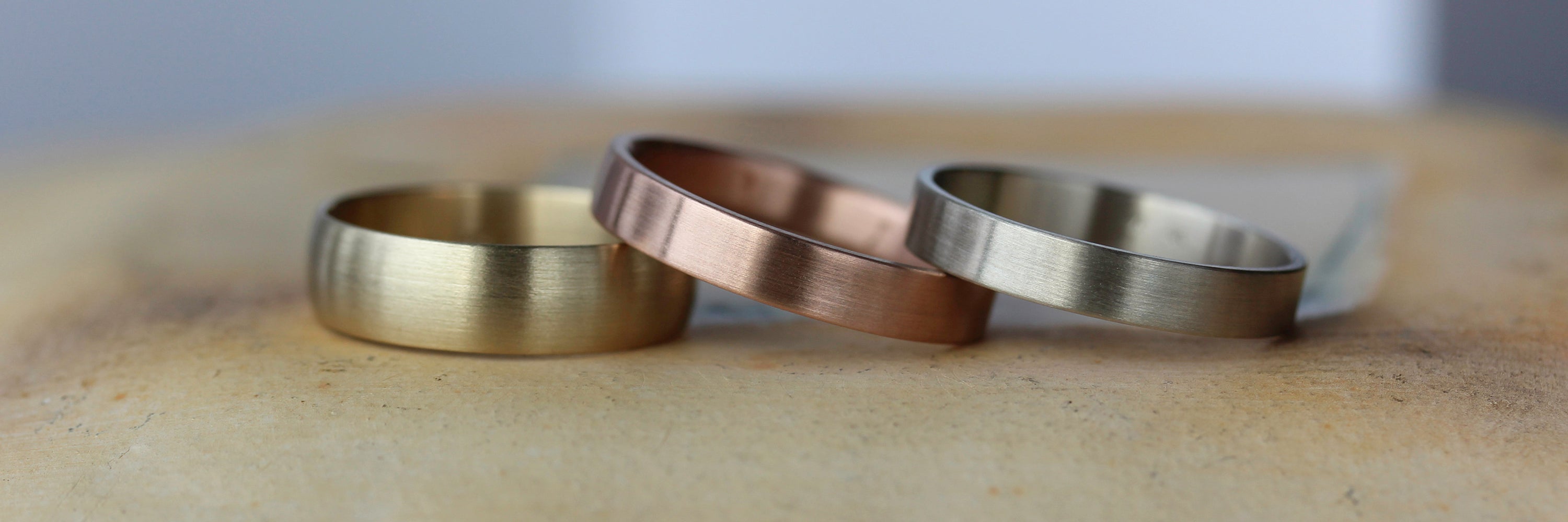 Three wide wedding bands on a tan surface showcase different gold tones. From left to right, yellow, rose, and white gold.