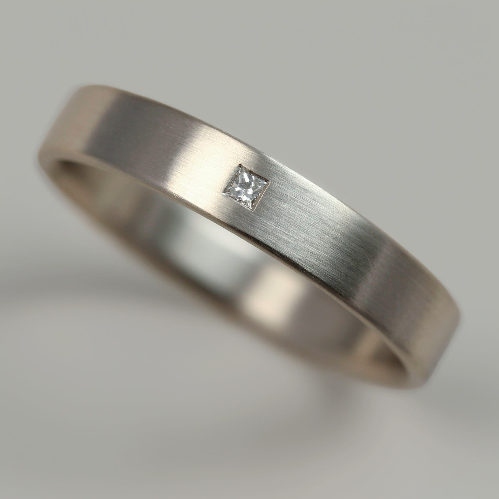 A plain smooth flat wedding band style with a flush set square diamond. The band has a matte white metal appearance.