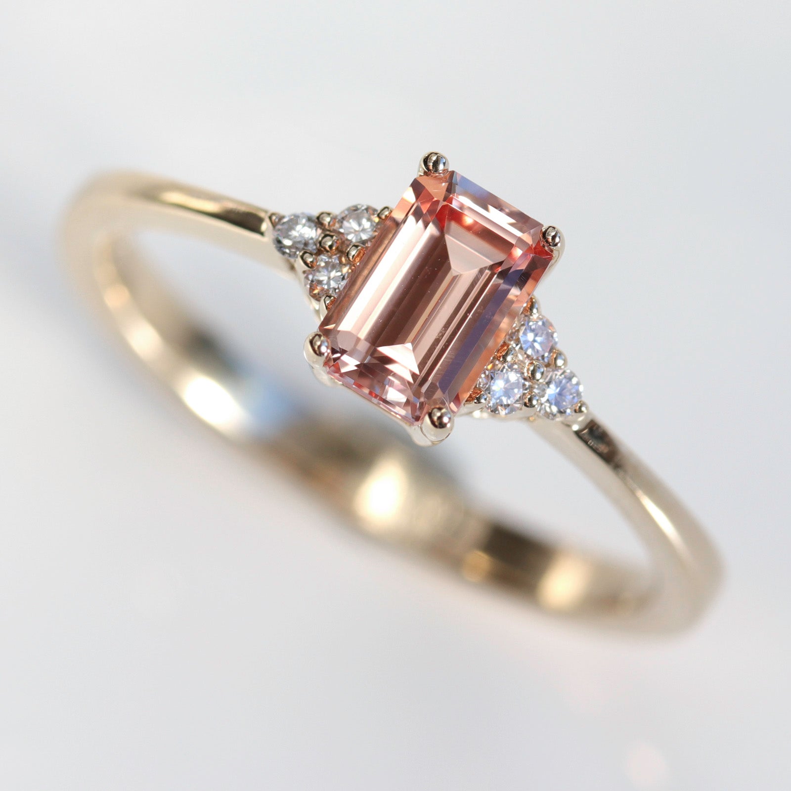 A seven stone cluster engagement ring style in shiny yellow gold. The ring has a peach champagne sapphire center stone in an emerald cut.