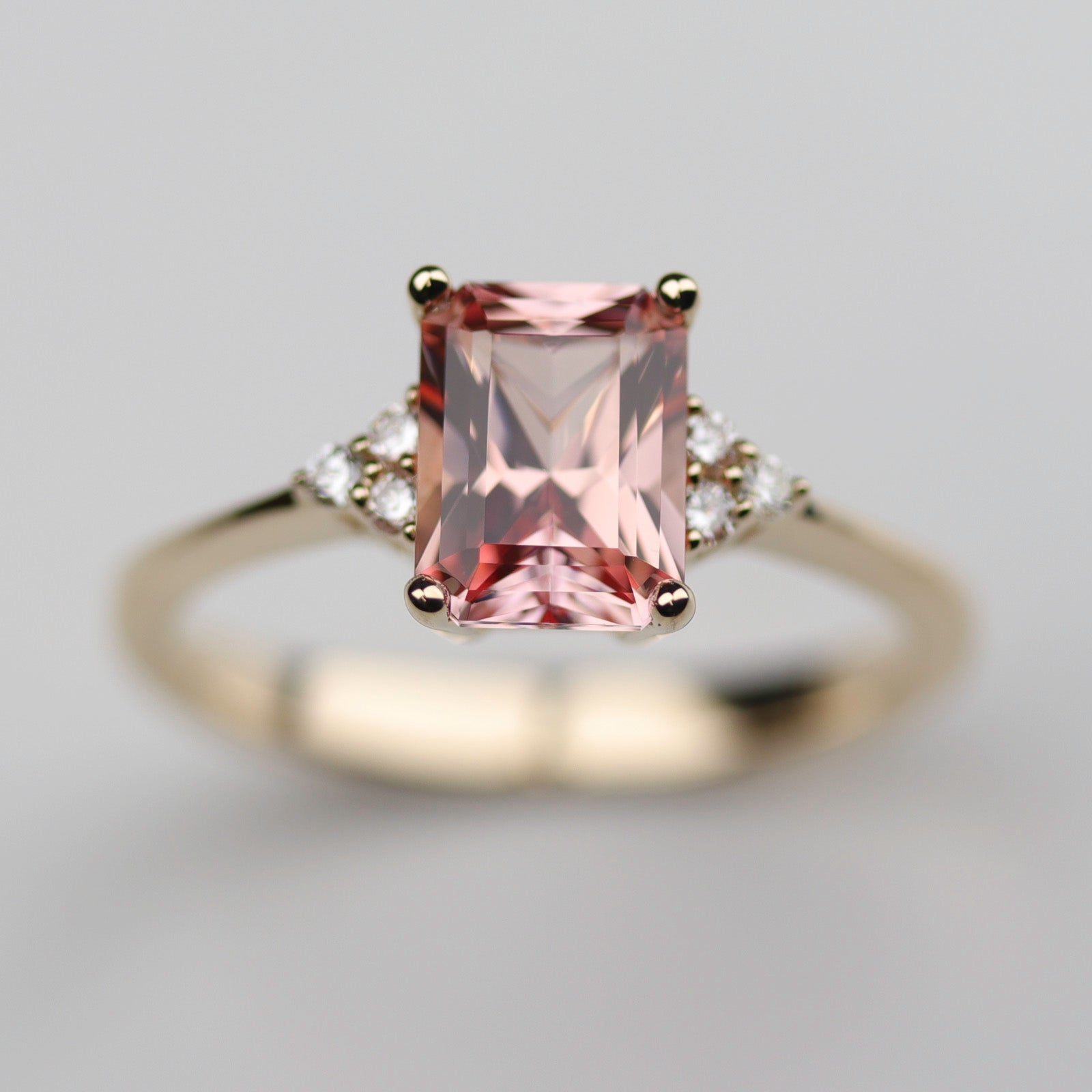 A seven stone cluster engagement ring style in shiny yellow gold. The ring has a peach champagne sapphire center stone in an emerald cut.
